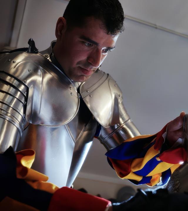 Ein Mitglied der Schweizer Garde trägt eine glänzende Rüstung und poliert ein Schild, während im Hintergrund Hellebarden zu sehen sind.