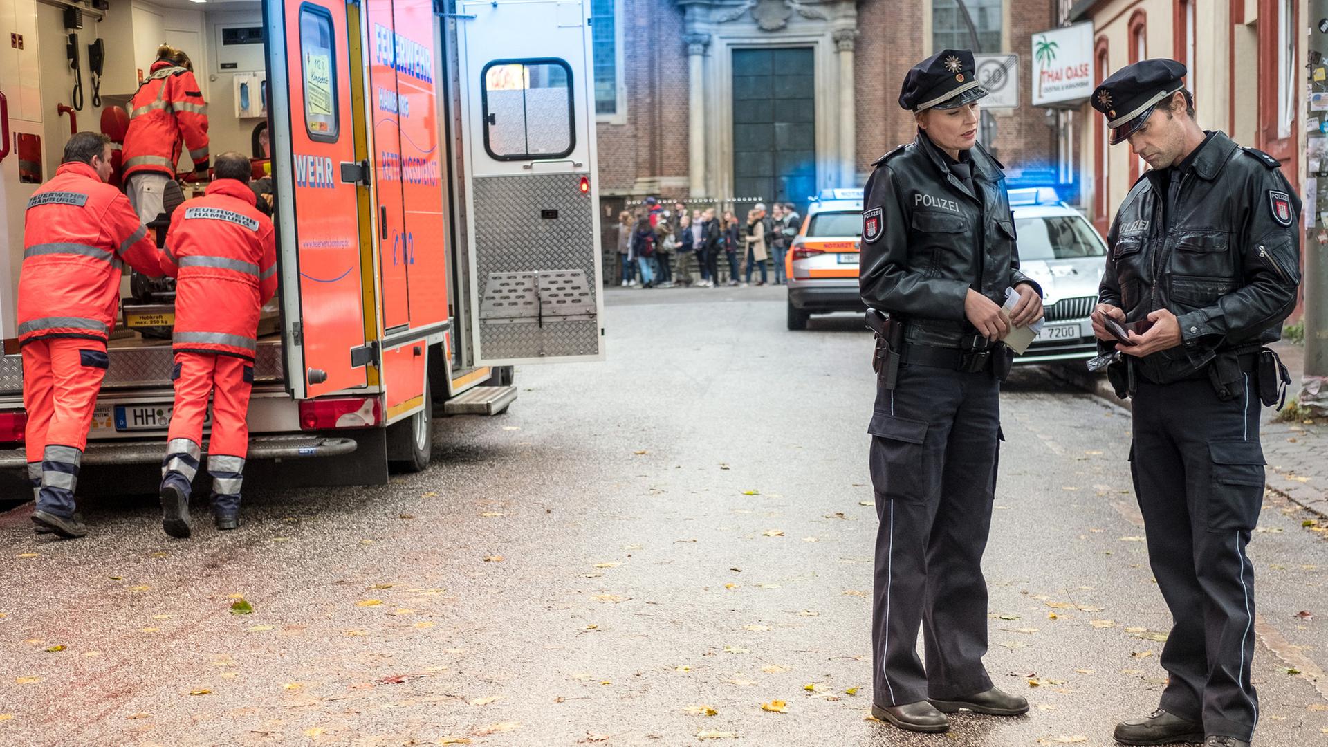 "Notruf Hafenkante - Die Sex-Party": Die Polizisten Melanie Hansen (Sanna Englund) und Mattes Seeler (Matthias Schloo) stehen nebeneinander auf einer Straße. Mattes hält einen Geldbeutel in den Händen und schaut auf den Ausweis, der sich darin befindet. Auf der anderen Straßenseite ein Notarztwagen. Zwei Rettungssanitäter (Komparsen) sind dabei eine Patiententrage in den Wagen zu schieben.