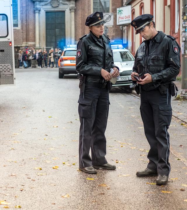"Notruf Hafenkante - Die Sex-Party": Die Polizisten Melanie Hansen (Sanna Englund) und Mattes Seeler (Matthias Schloo) stehen nebeneinander auf einer Straße. Mattes hält einen Geldbeutel in den Händen und schaut auf den Ausweis, der sich darin befindet. Auf der anderen Straßenseite ein Notarztwagen. Zwei Rettungssanitäter (Komparsen) sind dabei eine Patiententrage in den Wagen zu schieben.