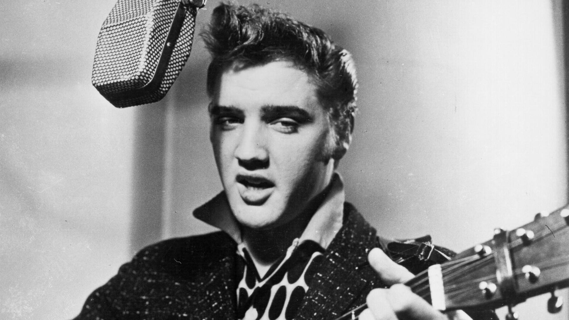 "Die sieben Leben des Elvis Presley": Schwarz-Weiß-Foto: Elvis Presley singt in ein Studiomikrofon und spielt dabei Gitarre.