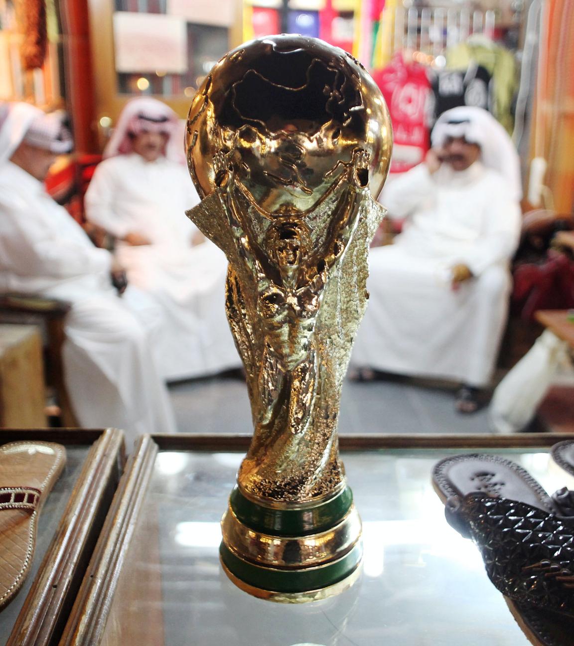 Im Vordergrund Nachbildung des FIFA WM-Pokals zwischen zwei Paar Schuhen. Im Hintergrund arabische Männer im Halbkreis sitzend.