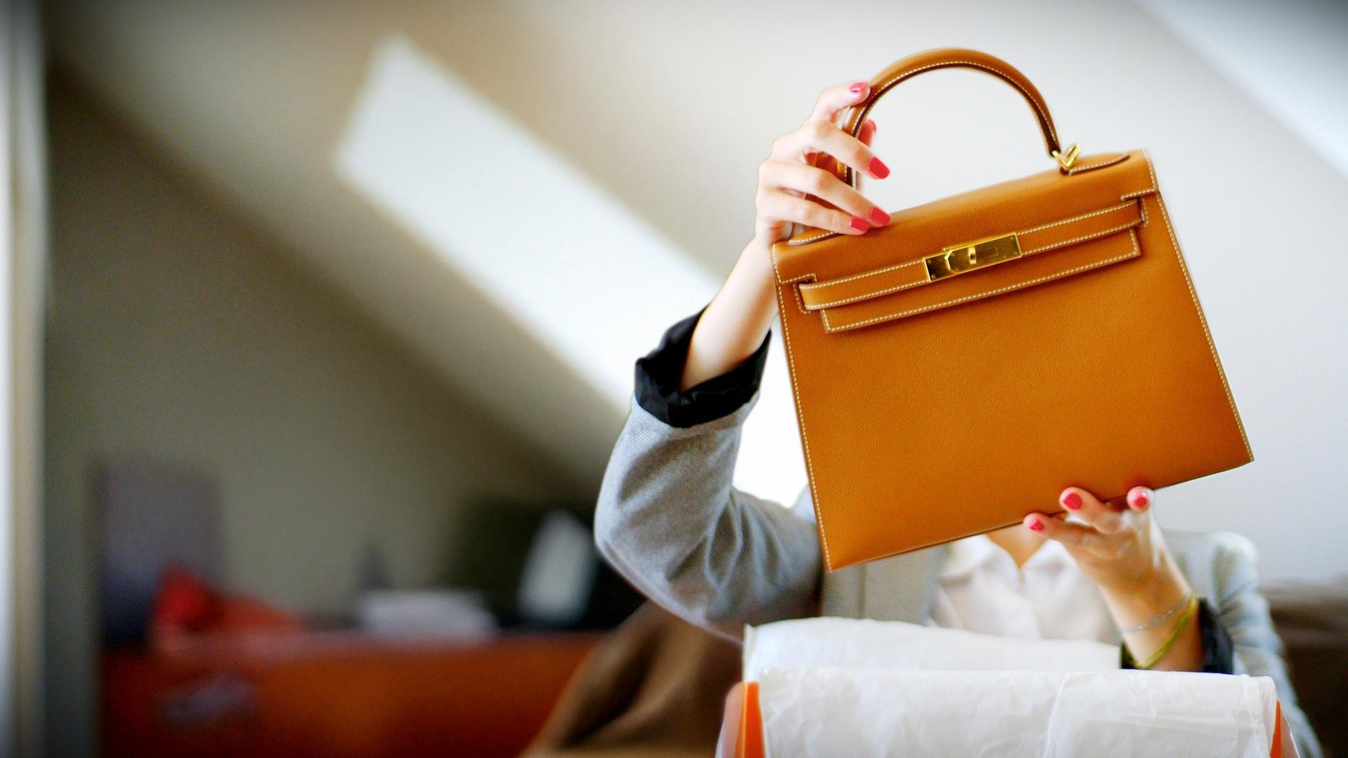 Frau packt stolz ihre neue Hermès Kelly aus und hält die Tasche vor ihrem Kopf in die Höhe.