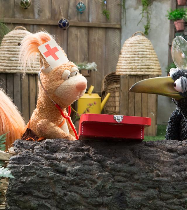 Eichhörnchen Ella und Rabe Rudi schauen sich fragend an. Sie tragen Häubchen und Spiegel auf dem Kopf, vor ihnen liegt ein kleiner Arztkoffer.