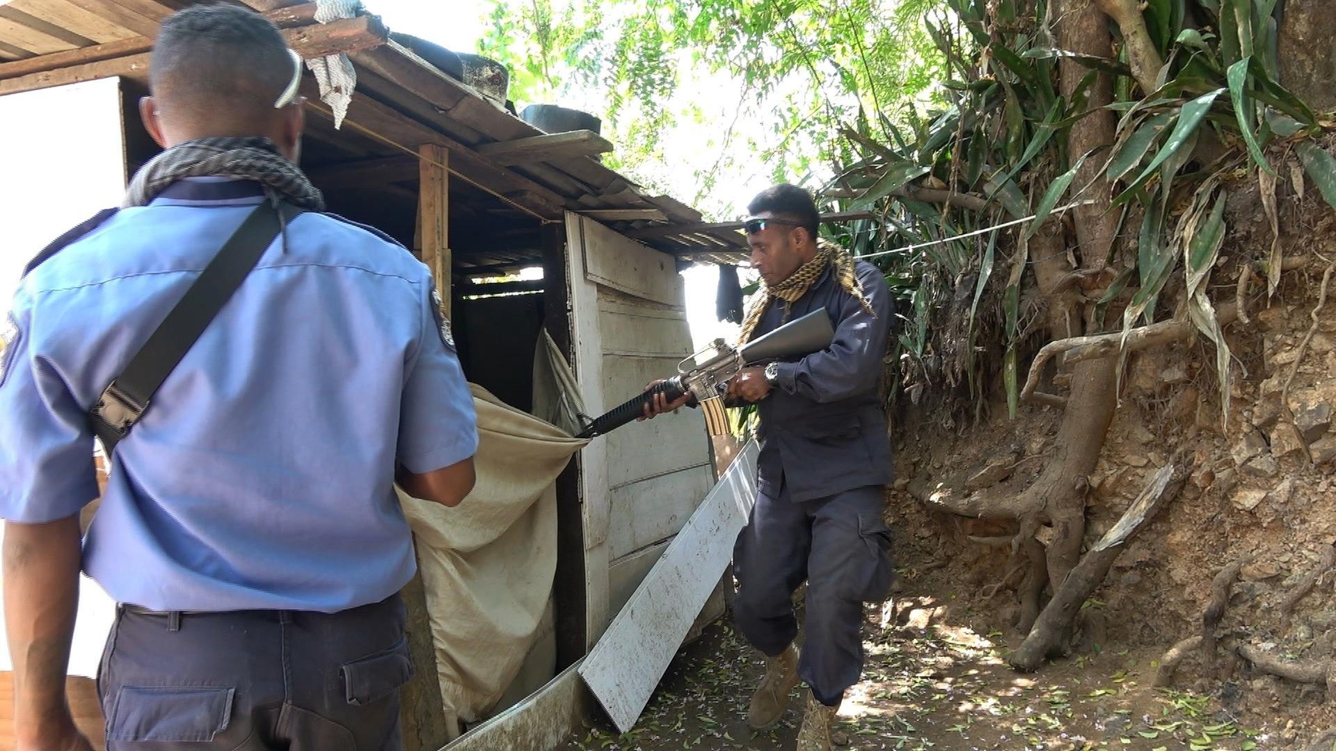 Zwei Männer stehen neben einer Holzhütte. Einer von ihnen hält eine Waffe und zielt in die Hütte hinein.