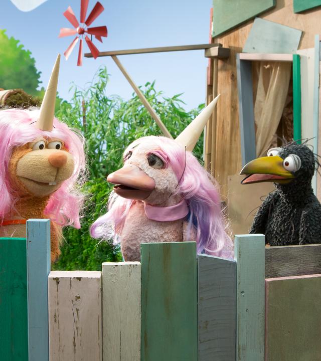 Rabe Rudi ist auf der Baumhaus-Plattform und schaut entsetzt auf Eichhörnchen Ella und Lina. Die beiden tragen rosa Perücken und Einhorn-Hörner auf den Köpfen und scheinen Rudi nicht zu bemerken.