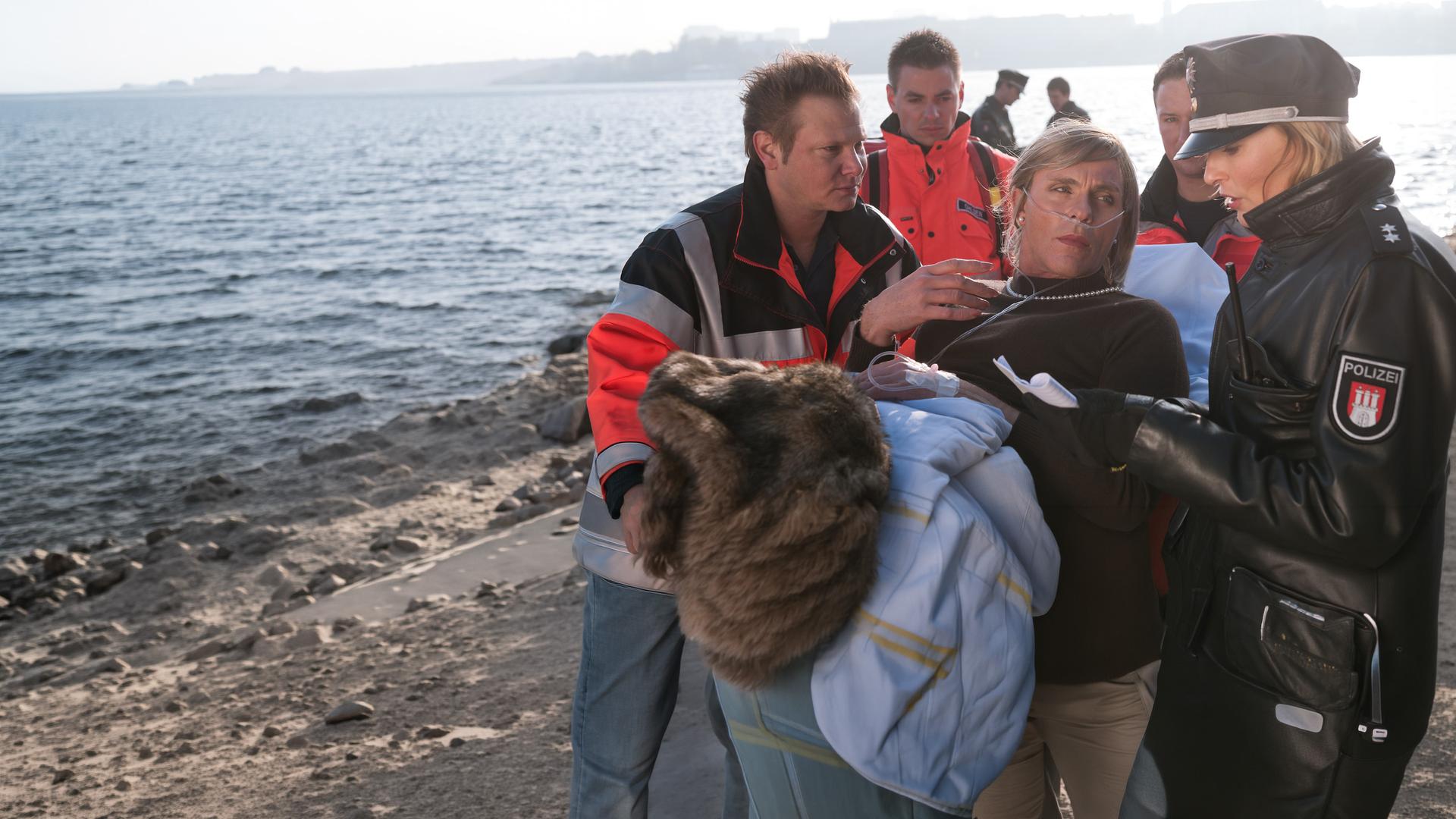 "Notruf Hafenkante - Die Tangotänzerin": Ramona Hellwig (Dirk Martens) wird, nachdem er im Ruderclub niedergeschlagen wurde, auf eine Trage gelegt. Dr. Haase (Fabian Harloff) kümmert sich um ihn. Melanie (Sanna Englund) befragt ihn währenddessen.