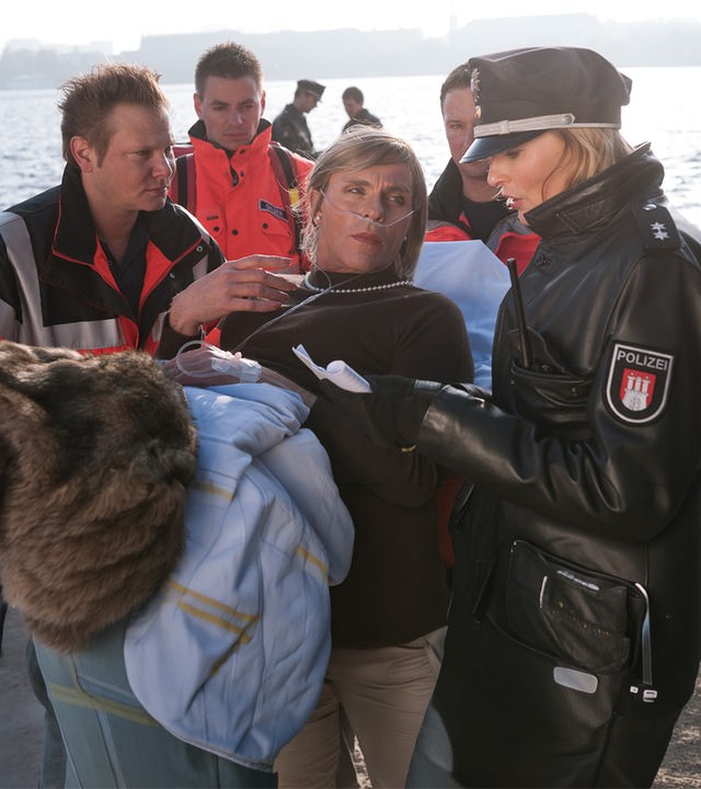 "Notruf Hafenkante - Die Tangotänzerin": Ramona Hellwig (Dirk Martens) wird, nachdem er im Ruderclub niedergeschlagen wurde, auf eine Trage gelegt. Dr. Haase (Fabian Harloff) kümmert sich um ihn. Melanie (Sanna Englund) befragt ihn währenddessen.