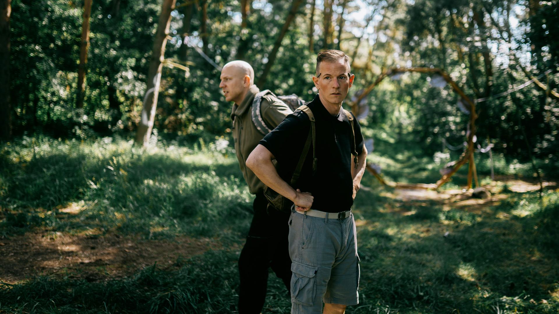 Zwei Personen befinden sich in einem bewaldeten Gebiet mit hohen Bäumen und üppigem Grün. Die Person im Vordergrund trägt ein schwarzes Shirt und graue Shorts, hat die Hände in die Hüften gestemmt und einen Rucksack auf dem Rücken. Die zweite Person steht etwas weiter hinten, trägt ein grünes Shirt und dunkle Hose und ebenfalls einen Rucksack. Sonnenlicht fällt durch die Bäume und erzeugt ein lebendiges Spiel aus Licht und Schatten auf dem Waldboden. Die Szene wirkt ruhig und natürlich und spielt sich offenbar tagsüber ab. 