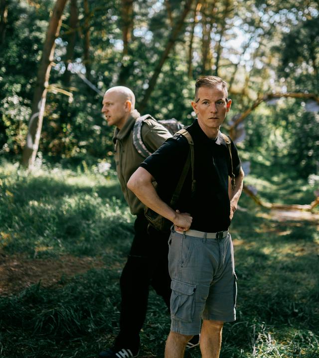 Zwei Personen befinden sich in einem bewaldeten Gebiet mit hohen Bäumen und üppigem Grün. Die Person im Vordergrund trägt ein schwarzes Shirt und graue Shorts, hat die Hände in die Hüften gestemmt und einen Rucksack auf dem Rücken. Die zweite Person steht etwas weiter hinten, trägt ein grünes Shirt und dunkle Hose und ebenfalls einen Rucksack. Sonnenlicht fällt durch die Bäume und erzeugt ein lebendiges Spiel aus Licht und Schatten auf dem Waldboden. Die Szene wirkt ruhig und natürlich und spielt sich offenbar tagsüber ab. 