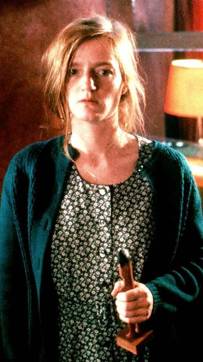 "Die tödliche Maria": Maria (Nina Petri) steht mitten in einem Raum, der durch Lampen warm erleuchtet ist. Sie starrt vor sich und hält in der linken Hand eine afrikanische Holzfigur.