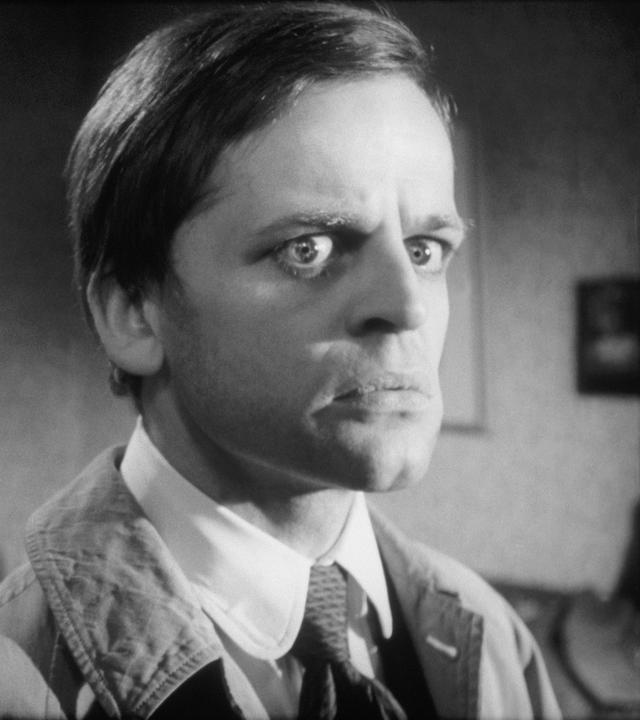 Spielszene in Schwarz-Weiß: Ein Mann im Anzug schaut beunruhigt, seine Augen sind weit aufgerissen. Er wird von einem weiteren Mann links von ihm beobachtet.