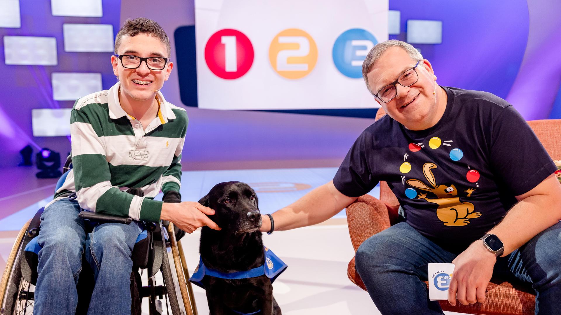 Paul sitzt im Rollstuhl neben Elton. Zwischen den beiden sitzt Assistenzhund Buena auf dem Boden und wird von Paul und Elton gestreichelt.