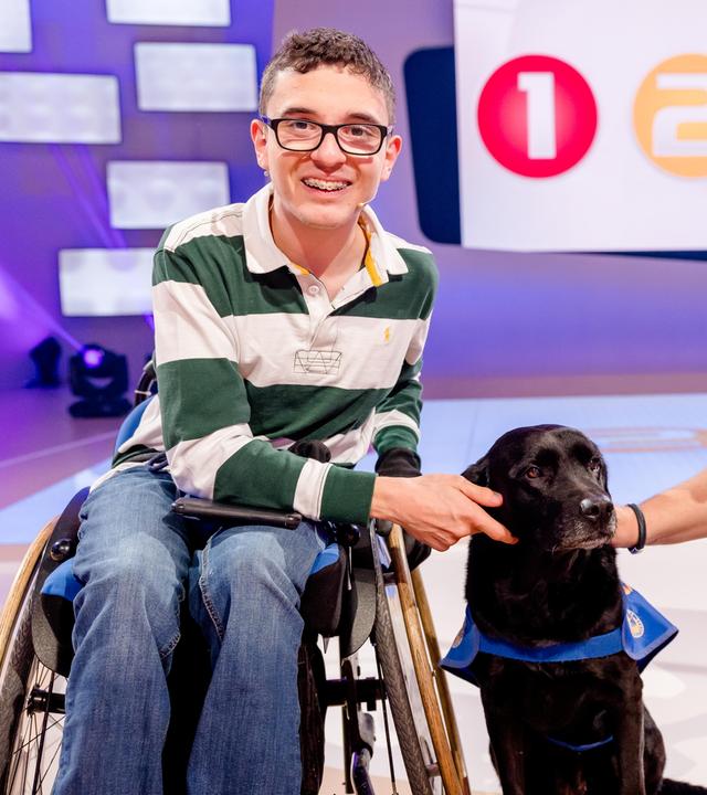 Paul sitzt im Rollstuhl neben Elton. Zwischen den beiden sitzt Assistenzhund Buena auf dem Boden und wird von Paul und Elton gestreichelt.