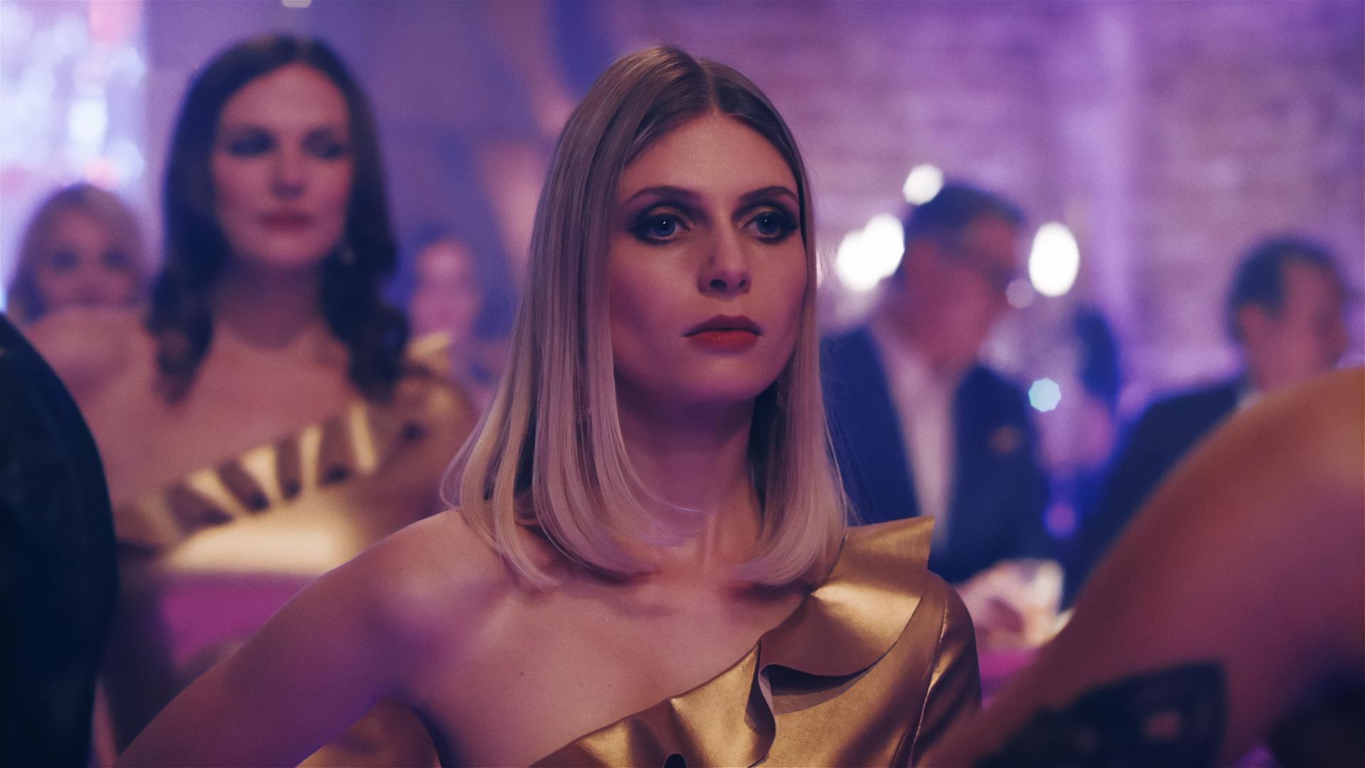 Ein blondes Model in einem goldenen Kleid. Im Hintergrund ein gemauertes Gewölbe und Partygäste. 