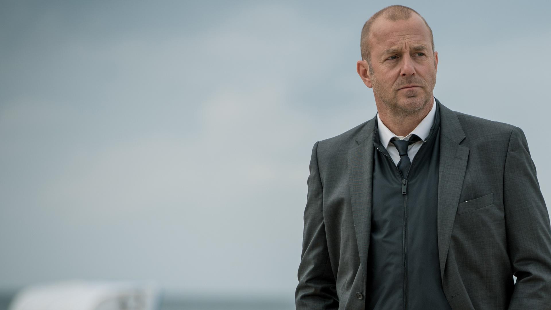 Kessler (Heino Ferch) steht am Strand. Im Hintergrund sind Strandkörbe zu erkennen.