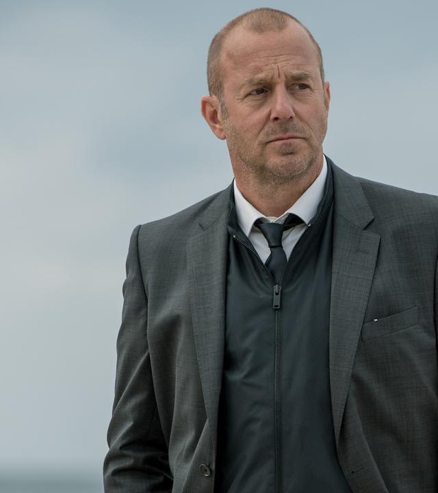 Kessler (Heino Ferch) steht am Strand. Im Hintergrund sind Strandkörbe zu erkennen.