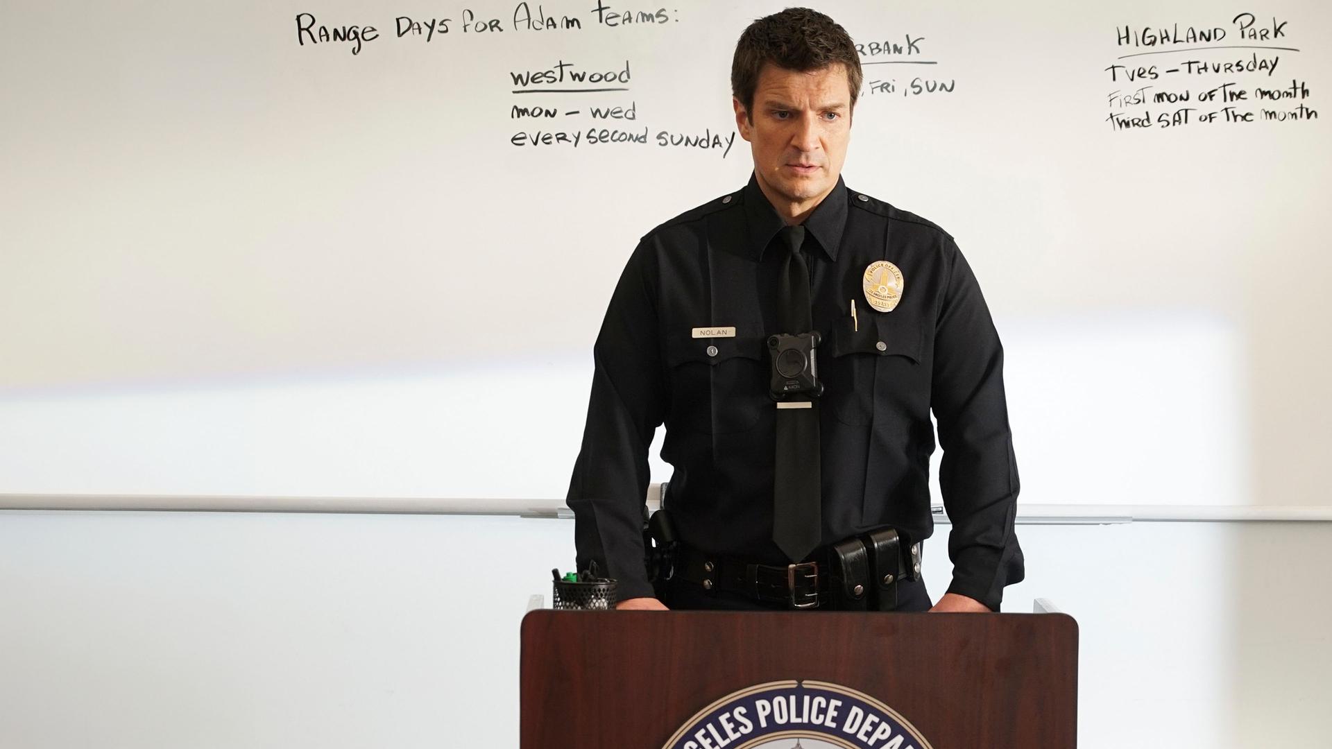 "The Rookie - Die Versuchung": Officer John Nolan (Nathan Fillion) steht vor einem Rednerpult. Und blickt in die Richtung seiner zuhörenden Kollegen. Im Hintergrund ein Whiteboard mit handschriftlichen Erörterungen.