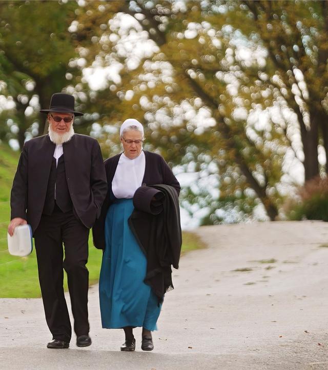 Ein älteres Paar in antiquierter Kleidung geht eine schmale Straße entlang.