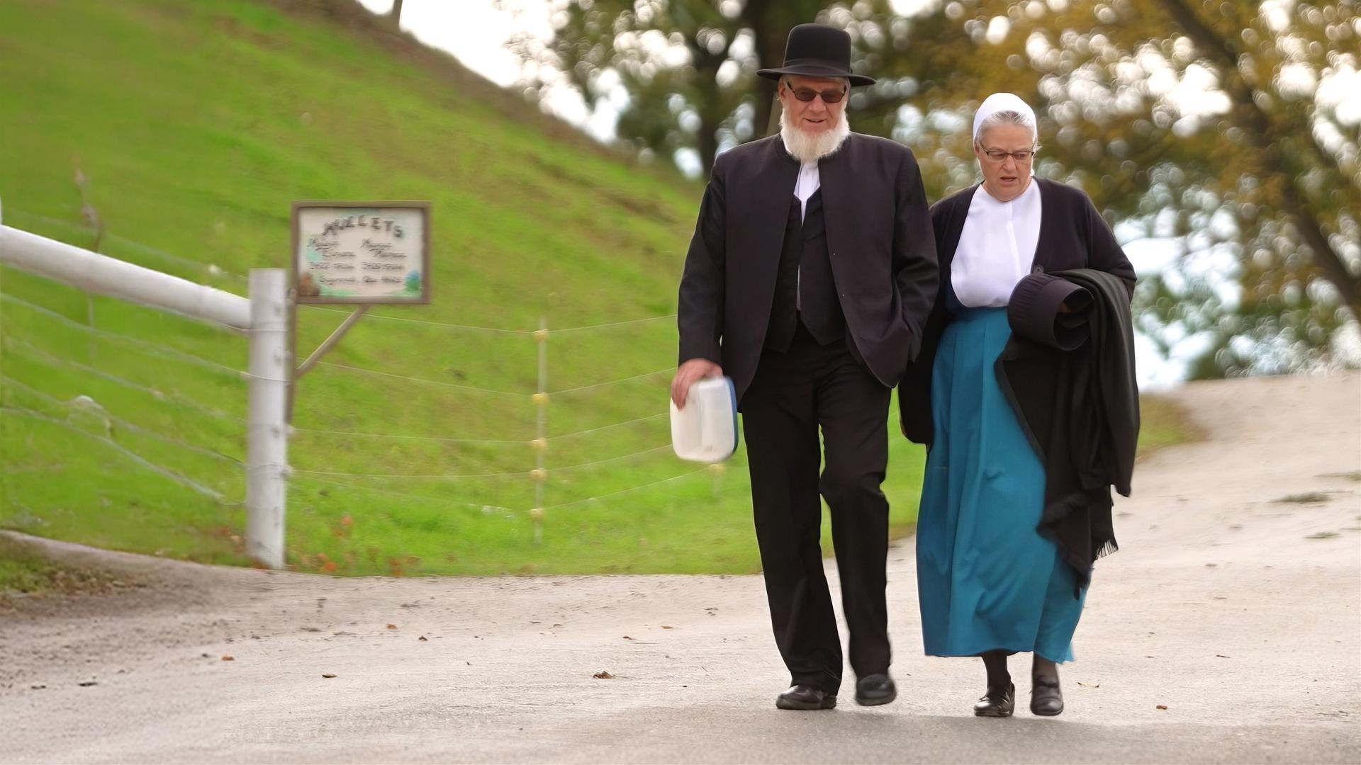 Ein älteres Paar in antiquierter Kleidung geht eine schmale Straße entlang.