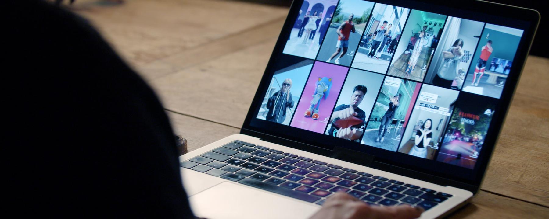 Ein aufgeklappter Laptop mit 12 Memeansichten, unscharf im Vordergrund eine Person von hinten, die vor dem Laptop sitzt.