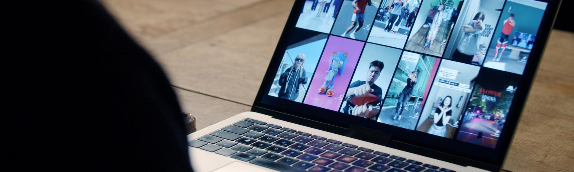 Ein aufgeklappter Laptop mit 12 Memeansichten, unscharf im Vordergrund eine Person von hinten, die vor dem Laptop sitzt.