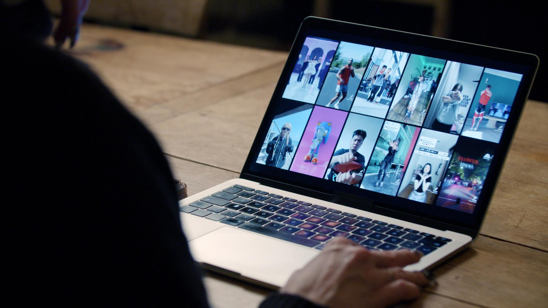 Ein aufgeklappter Laptop mit 12 Memeansichten, unscharf im Vordergrund eine Person von hinten, die vor dem Laptop sitzt.