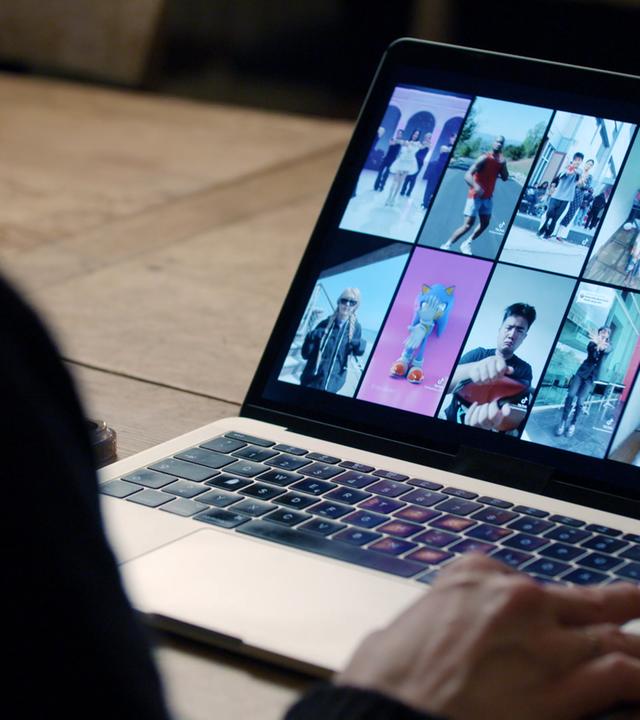 Ein aufgeklappter Laptop mit 12 Memeansichten, unscharf im Vordergrund eine Person von hinten, die vor dem Laptop sitzt.