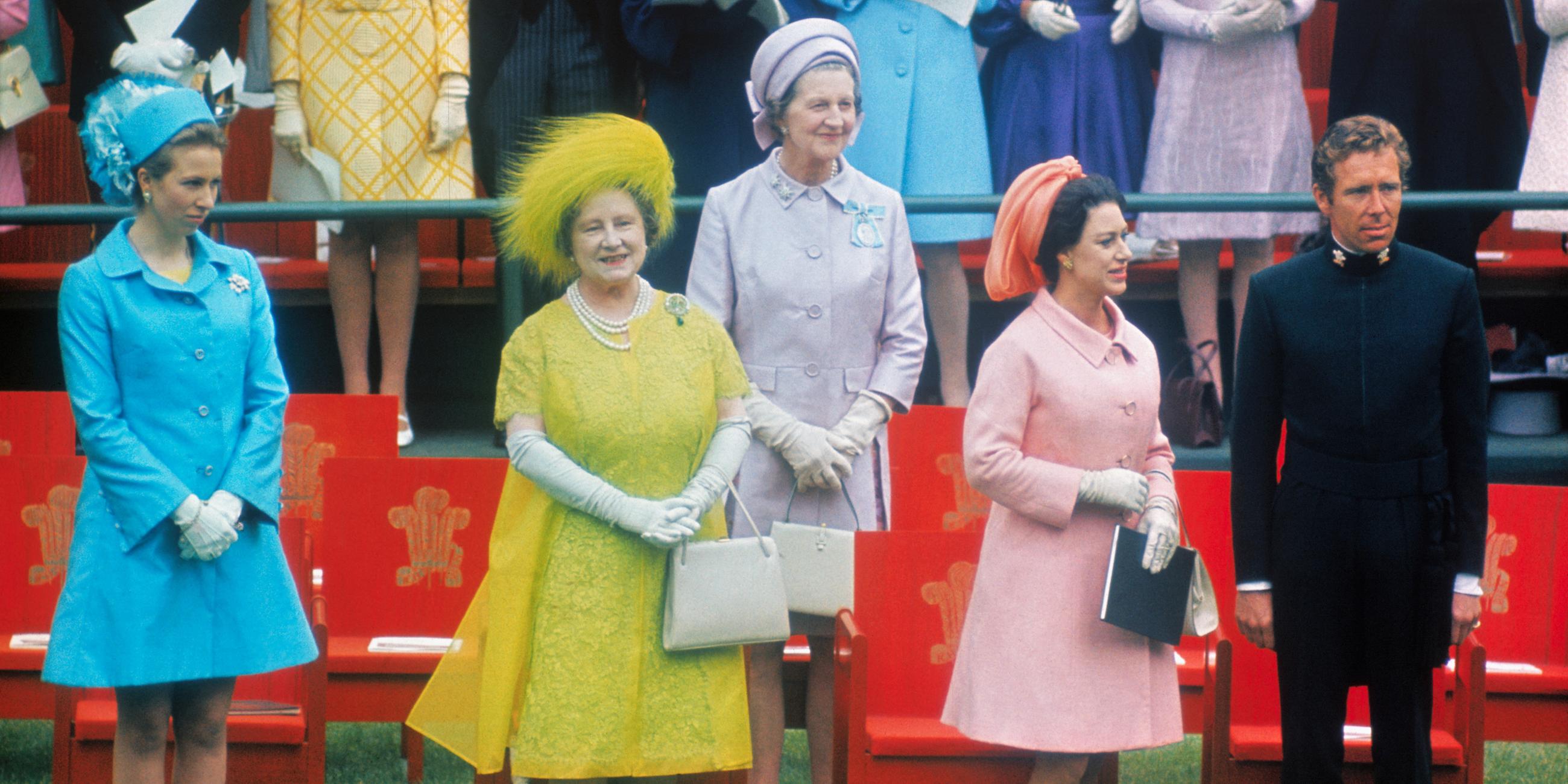  Elizabeth, die Queen Mum, steht in einem gelben Kleid zwischen anderen Frauen mit bunten Kleidern und Hüten und einem Mann in Uniform vor roten Stühlen.