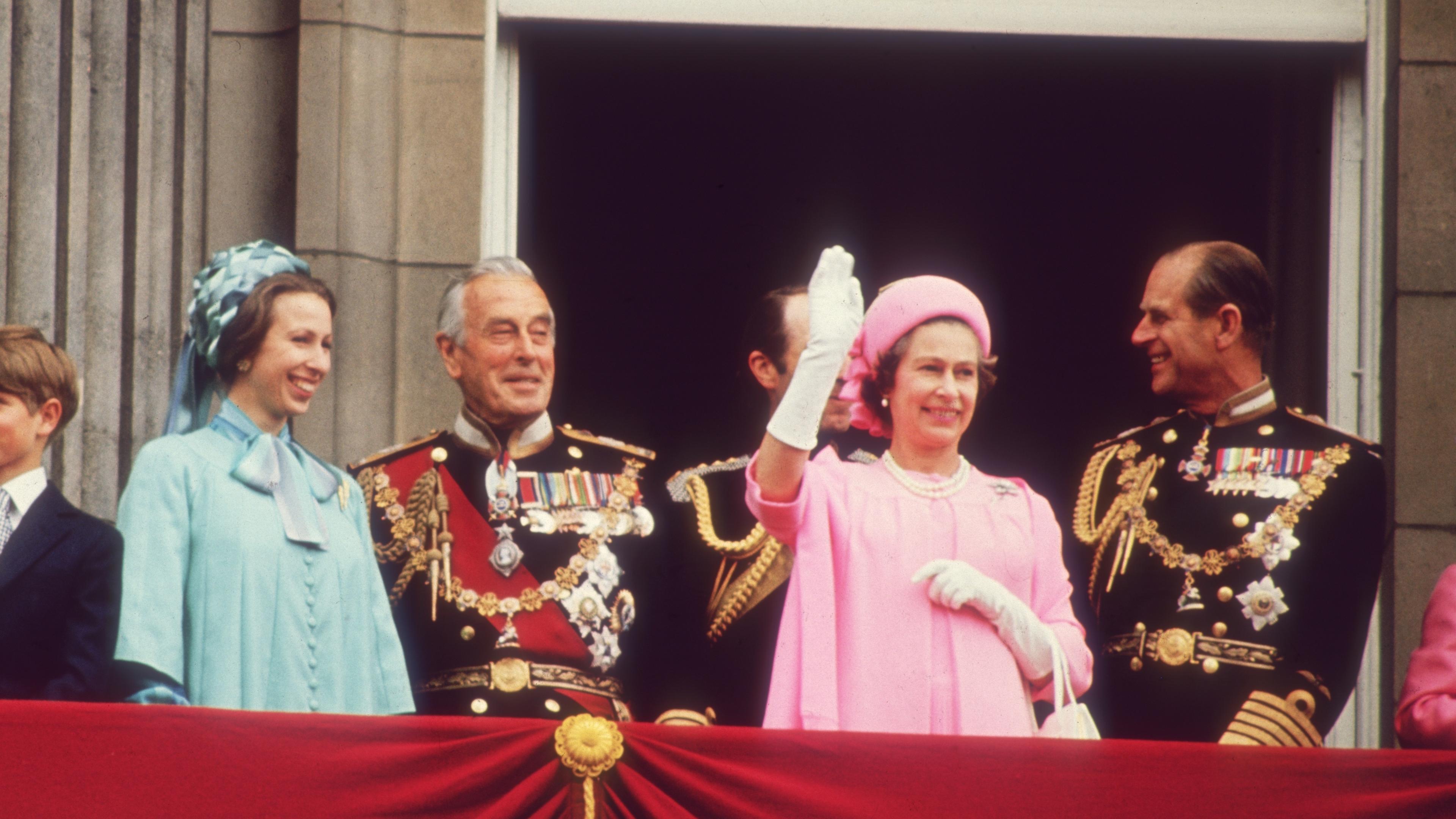  An einem Balkon, der mit einem roten Stoff dekoriert ist, stehen mehrere lächelnde Personen. Die Königin im rosafarbenen Kostüm grüßt mit der Hand.