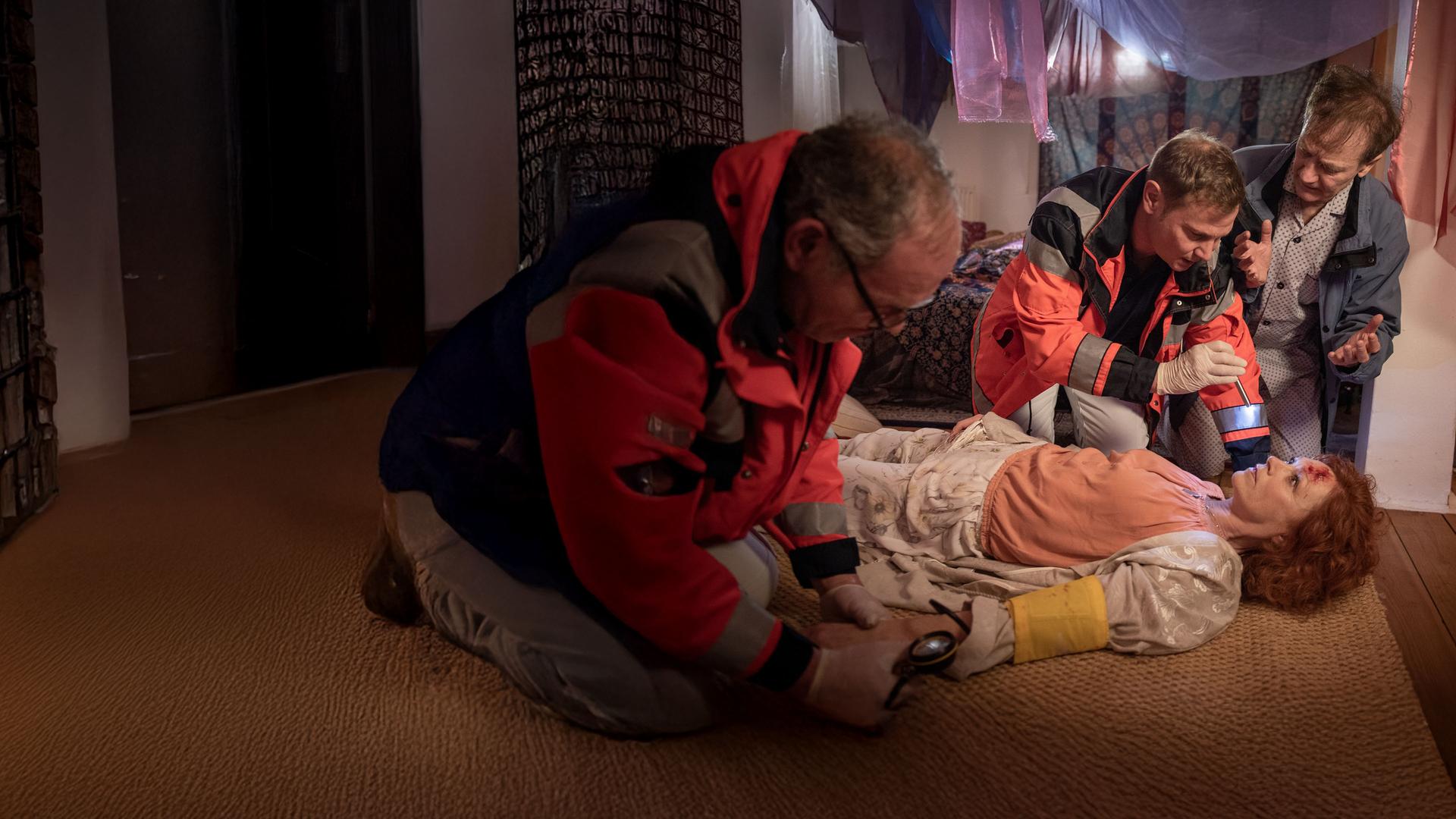 "Notruf Hafenkante - Die zauberhafte Adinda": Adinda Weyrauch (Heike Trinker) liegt auf dem Rücken auf dem Fußboden. Sie hat eine Verletzung an der Stirn. Dr. Haase (Fabian Harloff) leuchtet ihr mit einer Pupillenleuchte in die Augen. Herr Paus (Arnfried Lerche) hockt gestikulierend dicht neben ihm. Ein Rettungssanitäter (Komparse) hat Adinda am linken Arm eine Manschette angelegt und misst ihren Blutdruck.