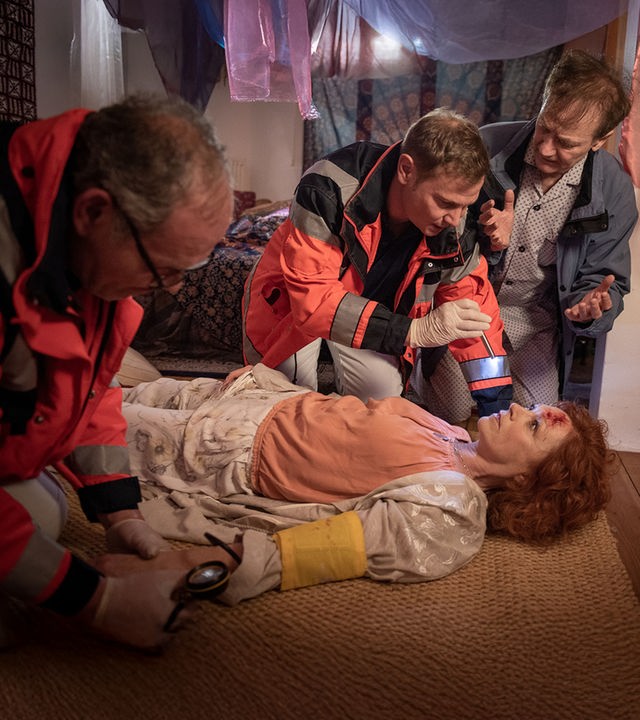 "Notruf Hafenkante - Die zauberhafte Adinda": Adinda Weyrauch (Heike Trinker) liegt auf dem Rücken auf dem Fußboden. Sie hat eine Verletzung an der Stirn. Dr. Haase (Fabian Harloff) leuchtet ihr mit einer Pupillenleuchte in die Augen. Herr Paus (Arnfried Lerche) hockt gestikulierend dicht neben ihm. Ein Rettungssanitäter (Komparse) hat Adinda am linken Arm eine Manschette angelegt und misst ihren Blutdruck.