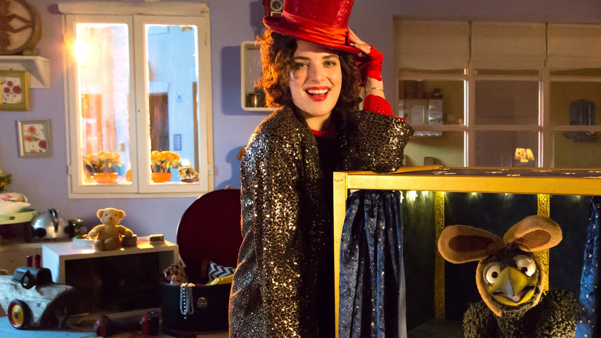 Siebenstein steht mit rotem Hut und Glitzermantel an einer offenen Kiste und lächelt Richtung Kamera. Rudi sitzt freudig in der Kiste und trägt Hasenohren. Im Hintergrund schaut Koffer zu.