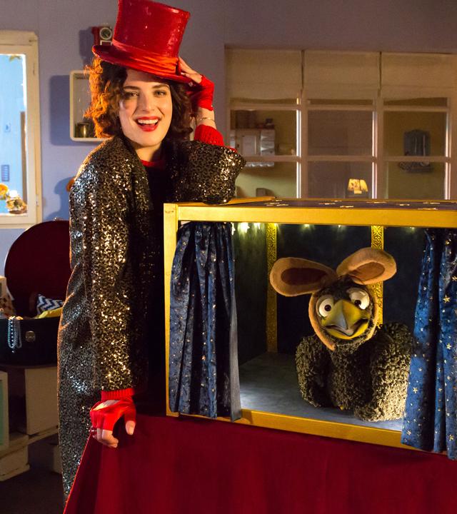 Siebenstein steht mit rotem Hut und Glitzermantel an einer offenen Kiste und lächelt Richtung Kamera. Rudi sitzt freudig in der Kiste und trägt Hasenohren. Im Hintergrund schaut Koffer zu.