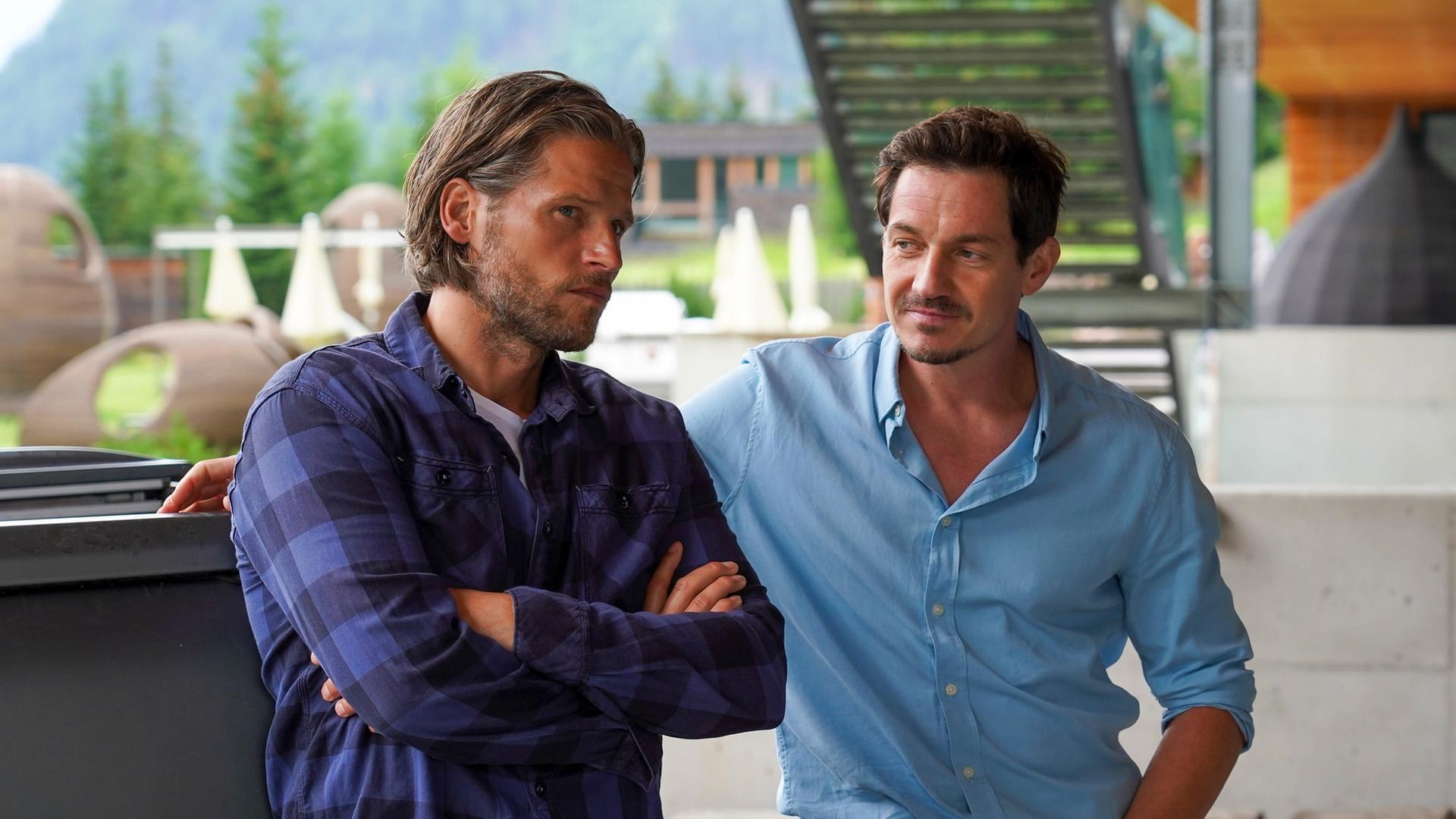 "Die Bergretter: Die Zeit, die bleibt": An ein Auto gelehnt steht Markus (Sebastian Ströbel) mit verschränkten Armen. Neben ihm steht Tobias (Markus Brandl) und blickt ihn fragend an.