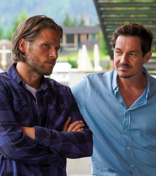 "Die Bergretter: Die Zeit, die bleibt": An ein Auto gelehnt steht Markus (Sebastian Ströbel) mit verschränkten Armen. Neben ihm steht Tobias (Markus Brandl) und blickt ihn fragend an.