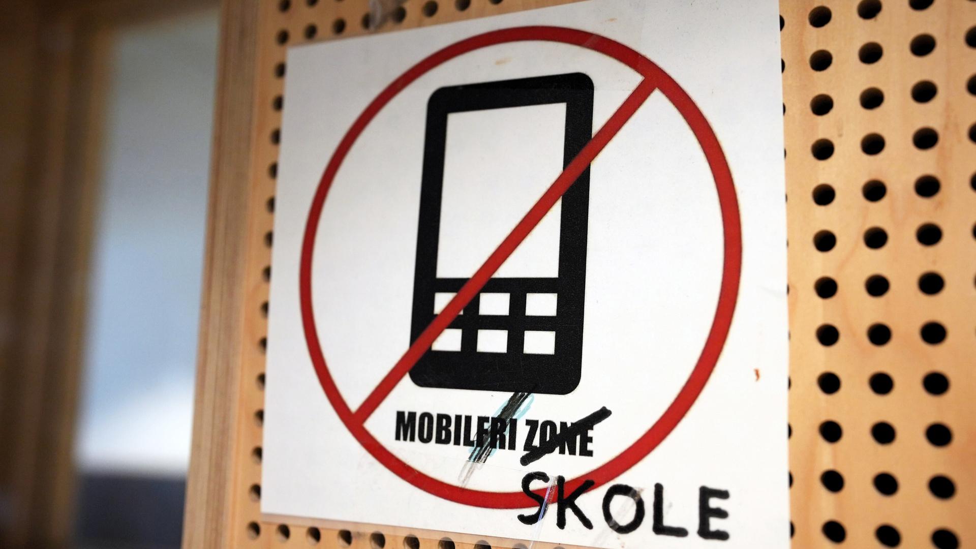 Das Bild zeigt ein Schild, das in einem öffentlichen oder schulischen Bereich angebracht ist. Auf dem Schild befindet sich ein durchgestrichenes Symbol eines Mobiltelefons, was darauf hinweist, dass Handynutzung an diesem Ort verboten ist. Der Text auf dem Schild besagt „MOBILFRI ZONE“, wobei das Wort „SKOLE“ zusätzlich in schwarzer Schrift unter dem ursprünglichen Text vermerkt ist, was auf einen Bezug zur Schule hindeutet. Der Hintergrund des Schildes ist ein perforiertes Holzbrett, das ebenfalls Teil der Umgebung sein könnte.   Das Bild thematisiert das Handyverbot in Schulen und könnte im Kontext der Diskussion über die Auswirkungen von Technologie auf die Jugend gesehen werden.
