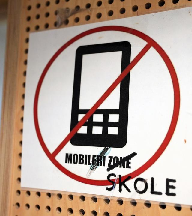 Das Bild zeigt ein Schild, das in einem öffentlichen oder schulischen Bereich angebracht ist. Auf dem Schild befindet sich ein durchgestrichenes Symbol eines Mobiltelefons, was darauf hinweist, dass Handynutzung an diesem Ort verboten ist. Der Text auf dem Schild besagt „MOBILFRI ZONE“, wobei das Wort „SKOLE“ zusätzlich in schwarzer Schrift unter dem ursprünglichen Text vermerkt ist, was auf einen Bezug zur Schule hindeutet. Der Hintergrund des Schildes ist ein perforiertes Holzbrett, das ebenfalls Teil der Umgebung sein könnte.   Das Bild thematisiert das Handyverbot in Schulen und könnte im Kontext der Diskussion über die Auswirkungen von Technologie auf die Jugend gesehen werden.