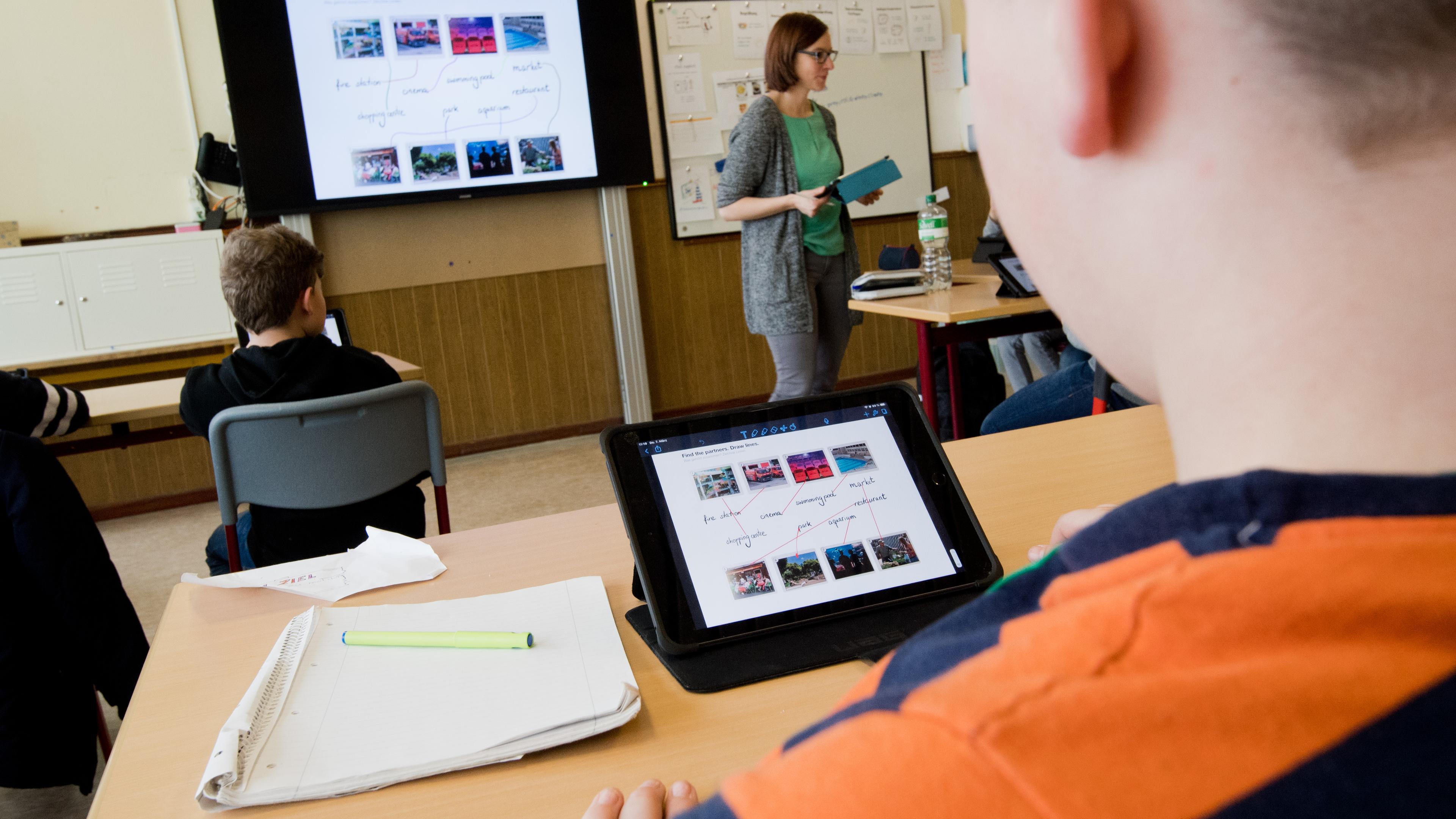 Ein Fünftklässler sitzt in einem Klassenzimmer vor einem iPad. Im Hintergrund ist eine digitale Tafel zu sehen.