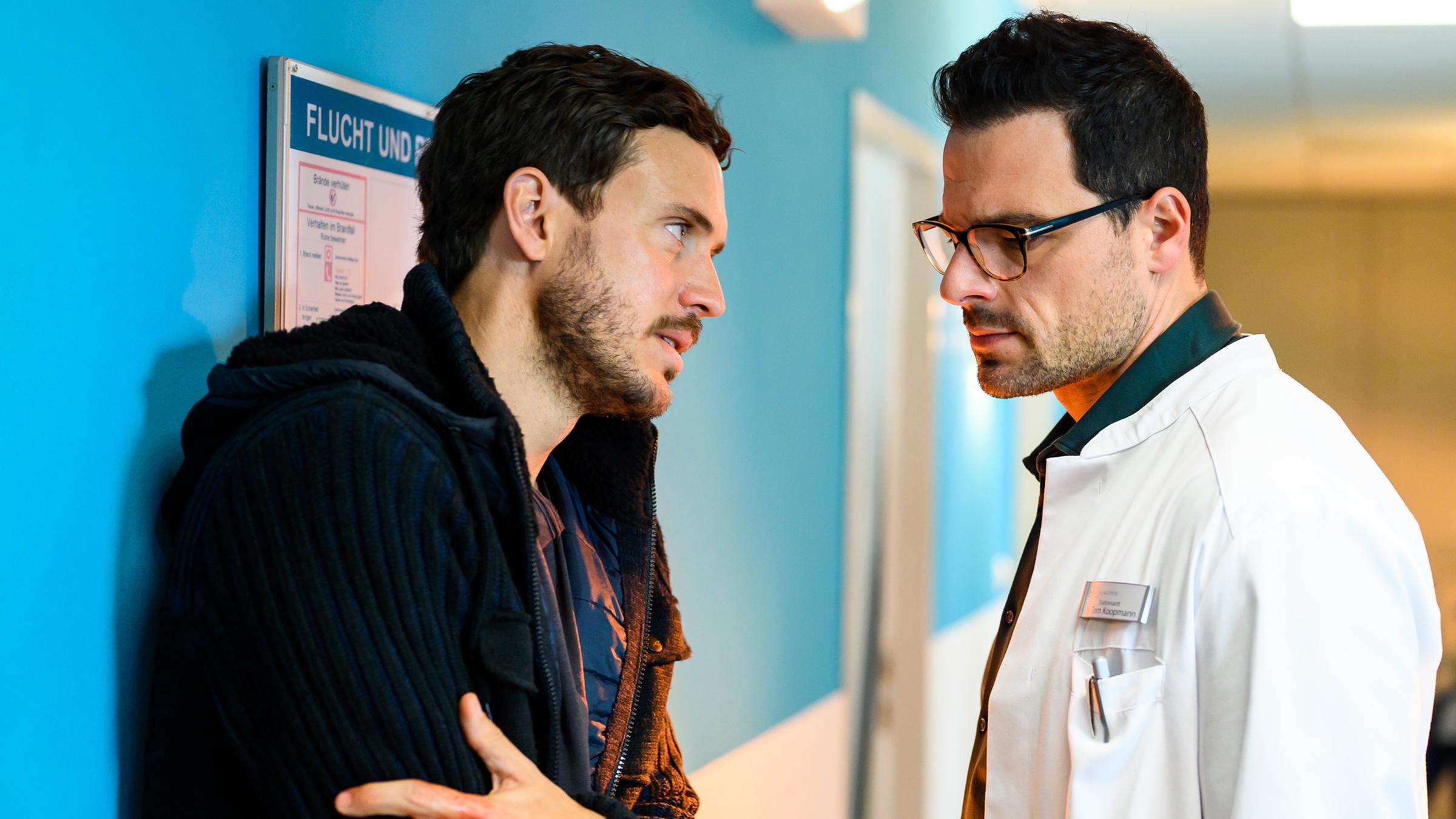 Mirko Koopmann (Tobias van Dieken) hat am rechten Arm eine Schussverletzung, auf die er seine linke Hand presst. Er steht im Flur der Karlsklinik mit hilfesuchendem Blick seinem Bruder Dr. Tom Koopmann (Tobias Licht) gegenüber und spricht mit ihm. Tom schaut mit erster Mine auf seine Verletzung.