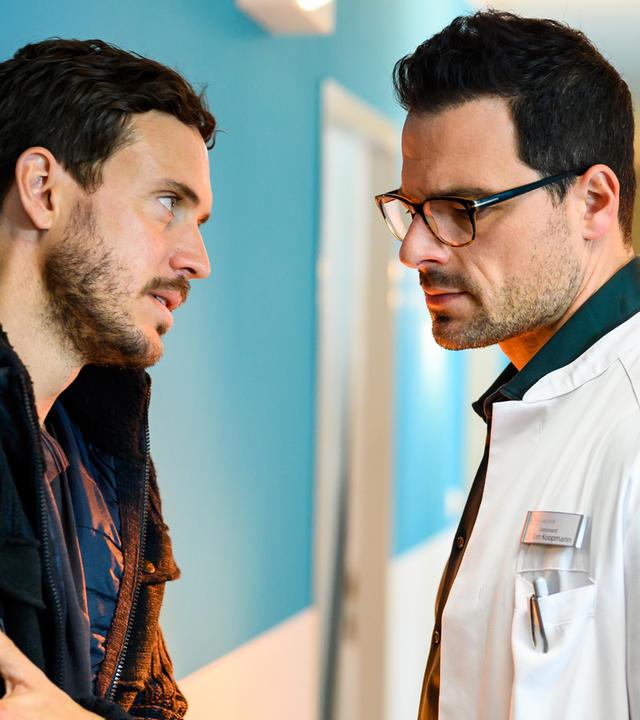 Mirko Koopmann (Tobias van Dieken) hat am rechten Arm eine Schussverletzung, auf die er seine linke Hand presst. Er steht im Flur der Karlsklinik mit hilfesuchendem Blick seinem Bruder Dr. Tom Koopmann (Tobias Licht) gegenüber und spricht mit ihm. Tom schaut mit erster Mine auf seine Verletzung.