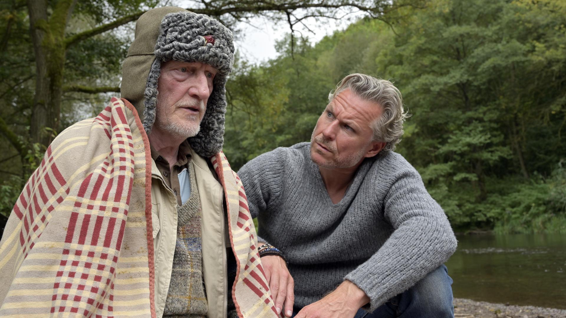 "Nächste Ausfahrt Glück - Juris Rückkehr": Will (Ernst Stötzner) sitzt mit einer Fellmütze auf dem Kopf und einer Decke um die Schultern an einem Flussufer, neben sich Juri (Dirk Borchardt)
