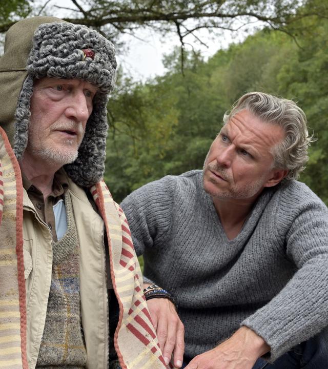 "Nächste Ausfahrt Glück - Juris Rückkehr": Will (Ernst Stötzner) sitzt mit einer Fellmütze auf dem Kopf und einer Decke um die Schultern an einem Flussufer, neben sich Juri (Dirk Borchardt)