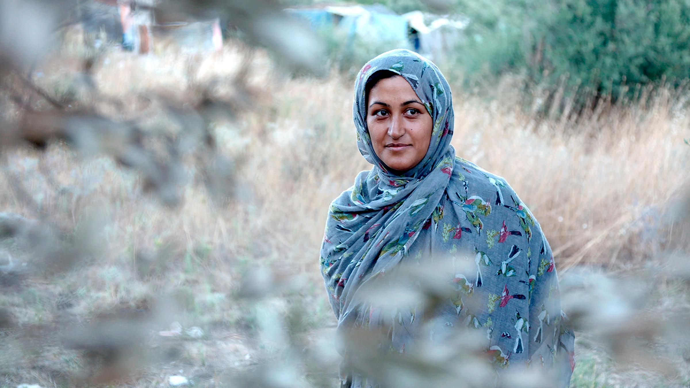 Nasim (Nasim Taijk) steht abseits von Zelten im Flüchtlingscamp hinter einem Baum. Sie trägt über ihrer Kleidung einen bunten Schleier und blickt lächelnd zu einer nicht im Bild zu sehenden Person.