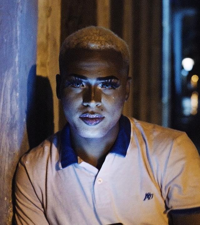Portraitbild eines schönen jungen Mannes mit kurzrasierten, blond gefärbten Haaren und geschminktem Gesicht.
