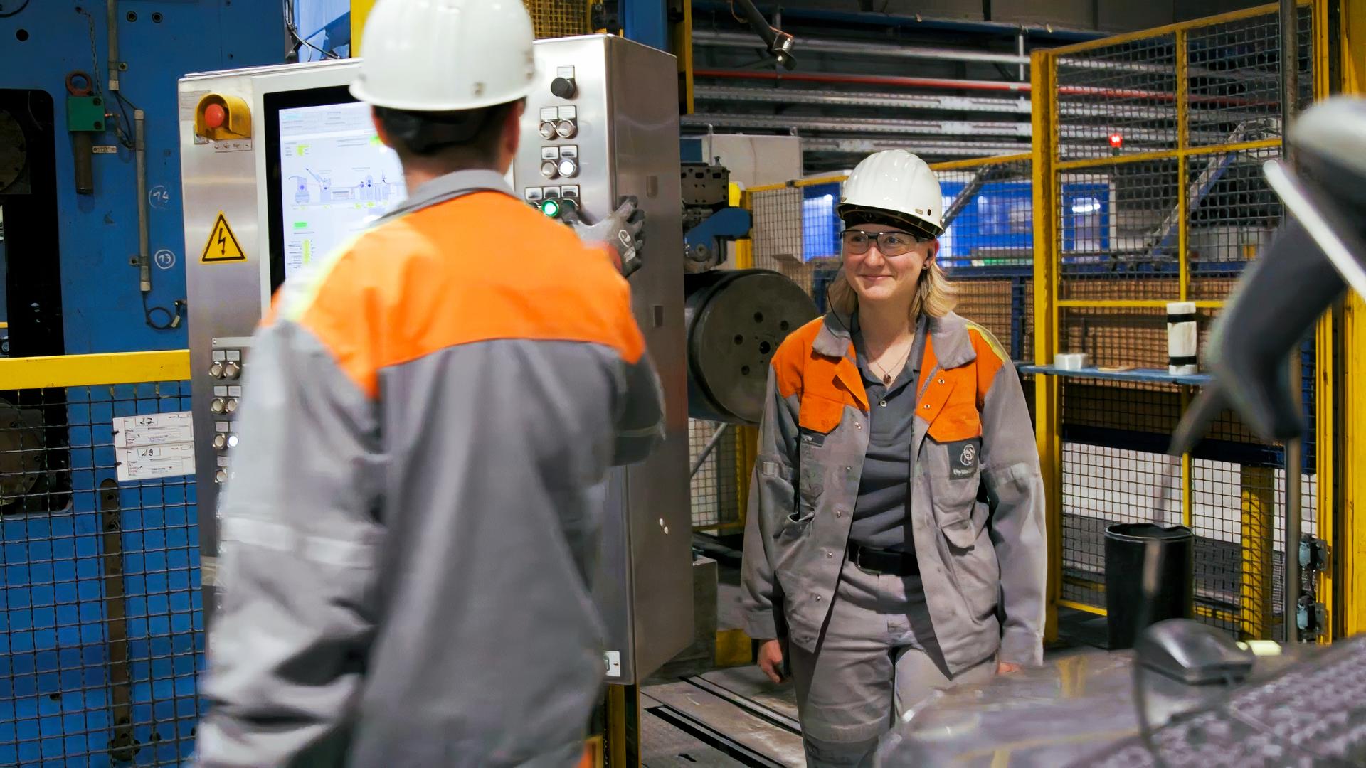 "DocuMe – Stahlarbeiterin": Eine junge Frau in Arbeitskleidung sowie mit Schutzhelm und Schutzbrille steht in einer Werkhalle rechts neben einem männlichen Kollegen, ebenfalls in Arbeitsmontur, der einen Kontrollmonitor im Blick hat.