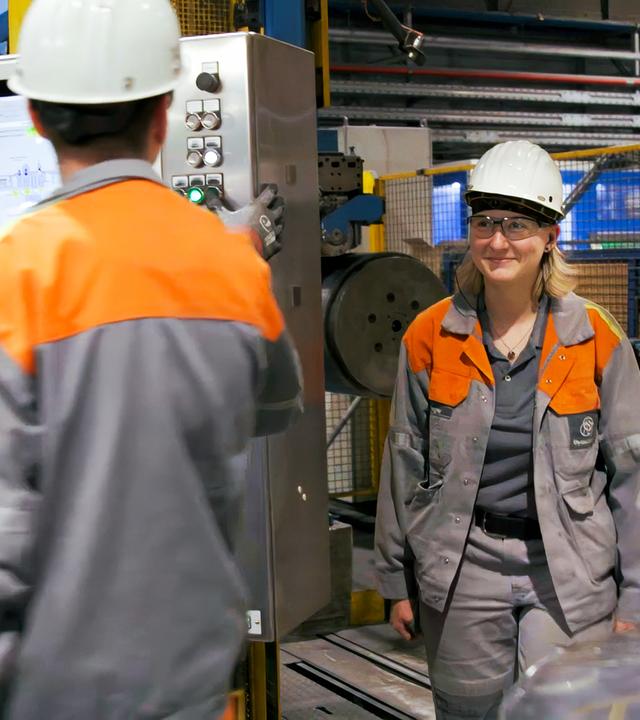 "DocuMe – Stahlarbeiterin": Eine junge Frau in Arbeitskleidung sowie mit Schutzhelm und Schutzbrille steht in einer Werkhalle rechts neben einem männlichen Kollegen, ebenfalls in Arbeitsmontur, der einen Kontrollmonitor im Blick hat.