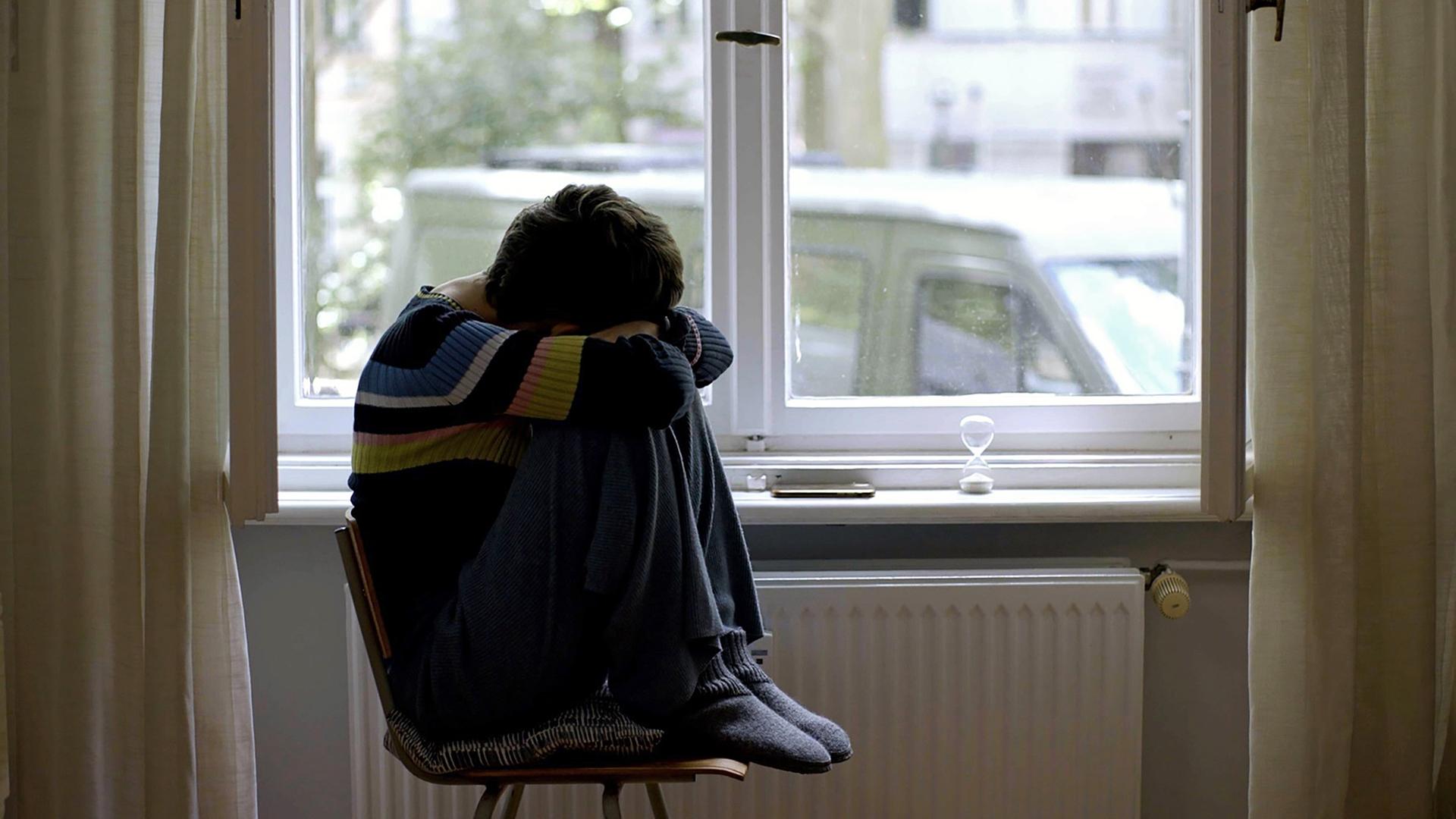 Junge Frau sitzt mit angezogenen Beinen auf einem Stuhl an einem Fenster.