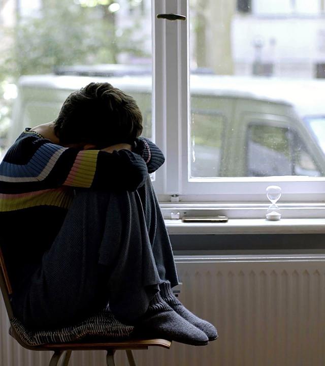 Junge Frau sitzt mit angezogenen Beinen auf einem Stuhl an einem Fenster.