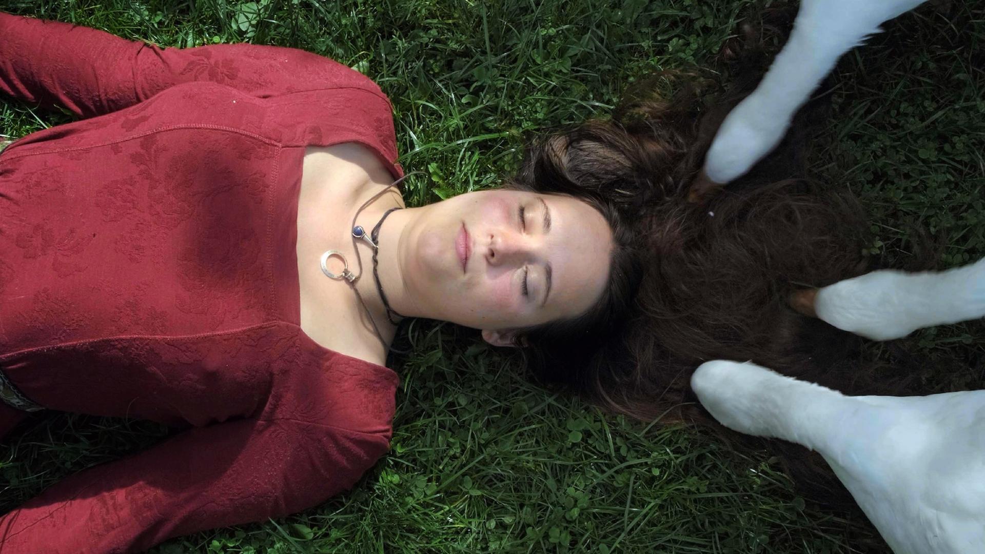 Eine Frau mit einem roten Oberteil liegt mit geschlossenen Augen auf eine Wiese. Weisse Kuhbeine stehen auf ihren langen brauen Haaren.