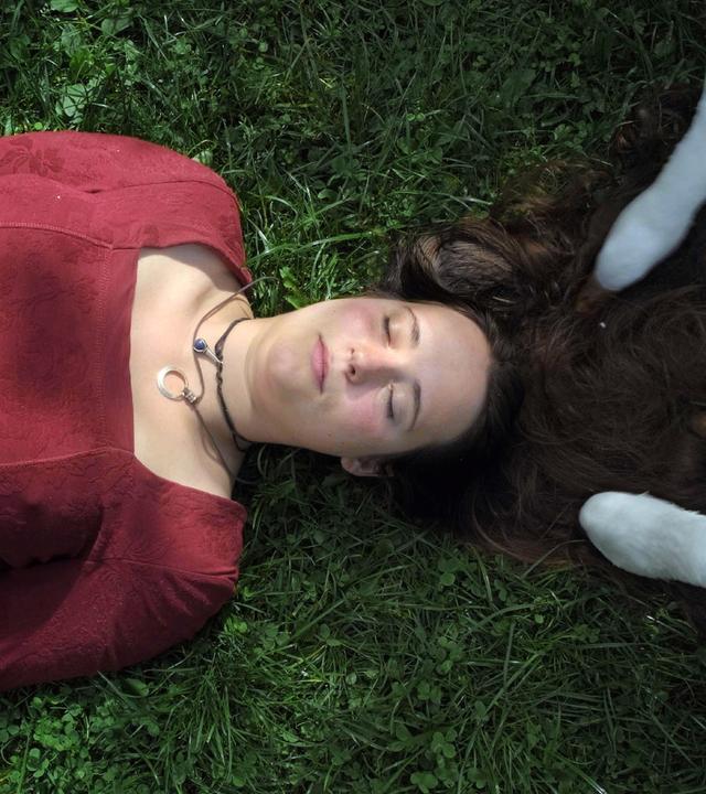 Eine Frau mit einem roten Oberteil liegt mit geschlossenen Augen auf eine Wiese. Weisse Kuhbeine stehen auf ihren langen brauen Haaren.